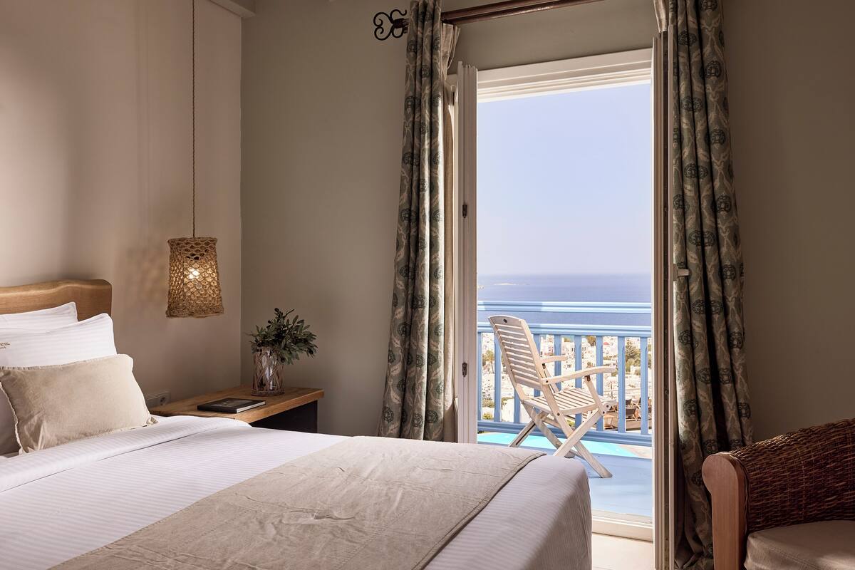 Een slaapkamer met een groot bed, een nachtkastje, een lamp en uitzicht op de oceaan door open gordijnen.