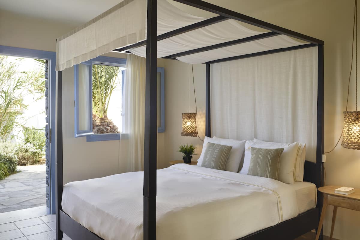 Een bed met hemelbed met witte lakens, een klein nachtkastje met een lamp, en uitzicht op de tuin door het raam.