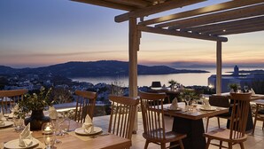 An outdoor dining area with wooden tables and chairs, set against a backdrop of a cityscape and water.