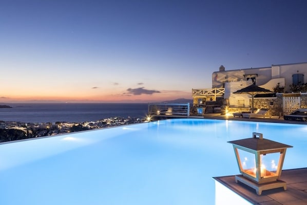 A poolside area with a view of the sea at dusk, a lantern, and a building with a balcony.