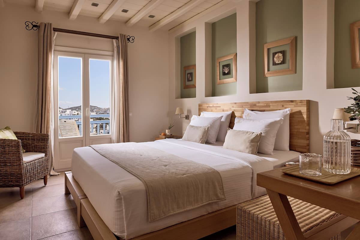Een slaapkamer met een groot bed, een houten hoofdbord, een nachtkastje, een rieten stoel en uitzicht op de natuur.