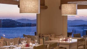A dining area with tables set for two, large windows with a view of the sea and hills, and hanging lamps.
