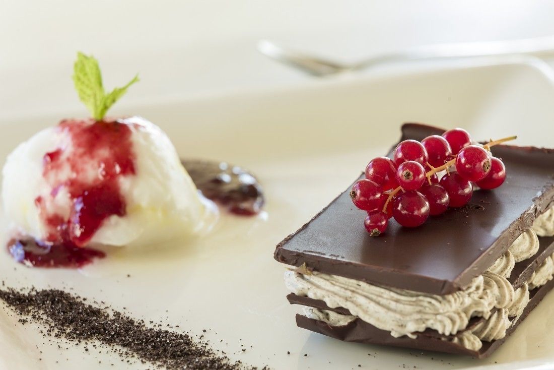 Un plato de postre con una bola de helado coronada con salsa de frutos rojos, un trozo de chocolate con capas de crema blanca y un racimo de bayas rojas.