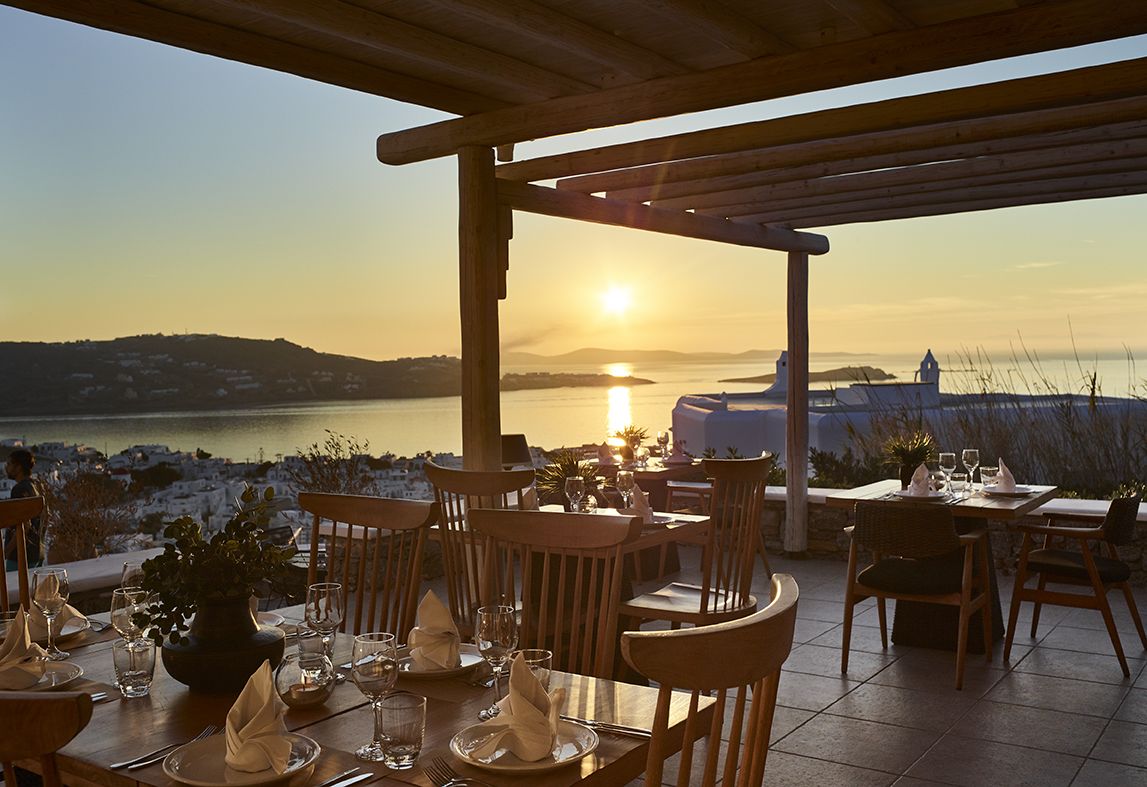 An outdoor dining area with wooden tables and chairs, set against a backdrop of a sunset over a body of water and hills.