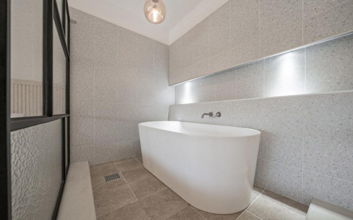 Ein modernes Badezimmer mit einer freistehenden Badewanne, gefliesten Wänden und einem sichtbaren Lüftungsgitter am Boden.