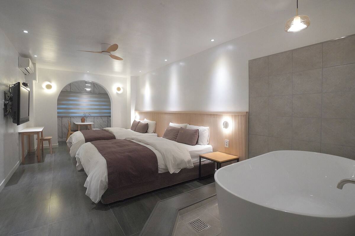 A modern bedroom with a large bed, a small table, a wall-mounted TV, and a bathtub.