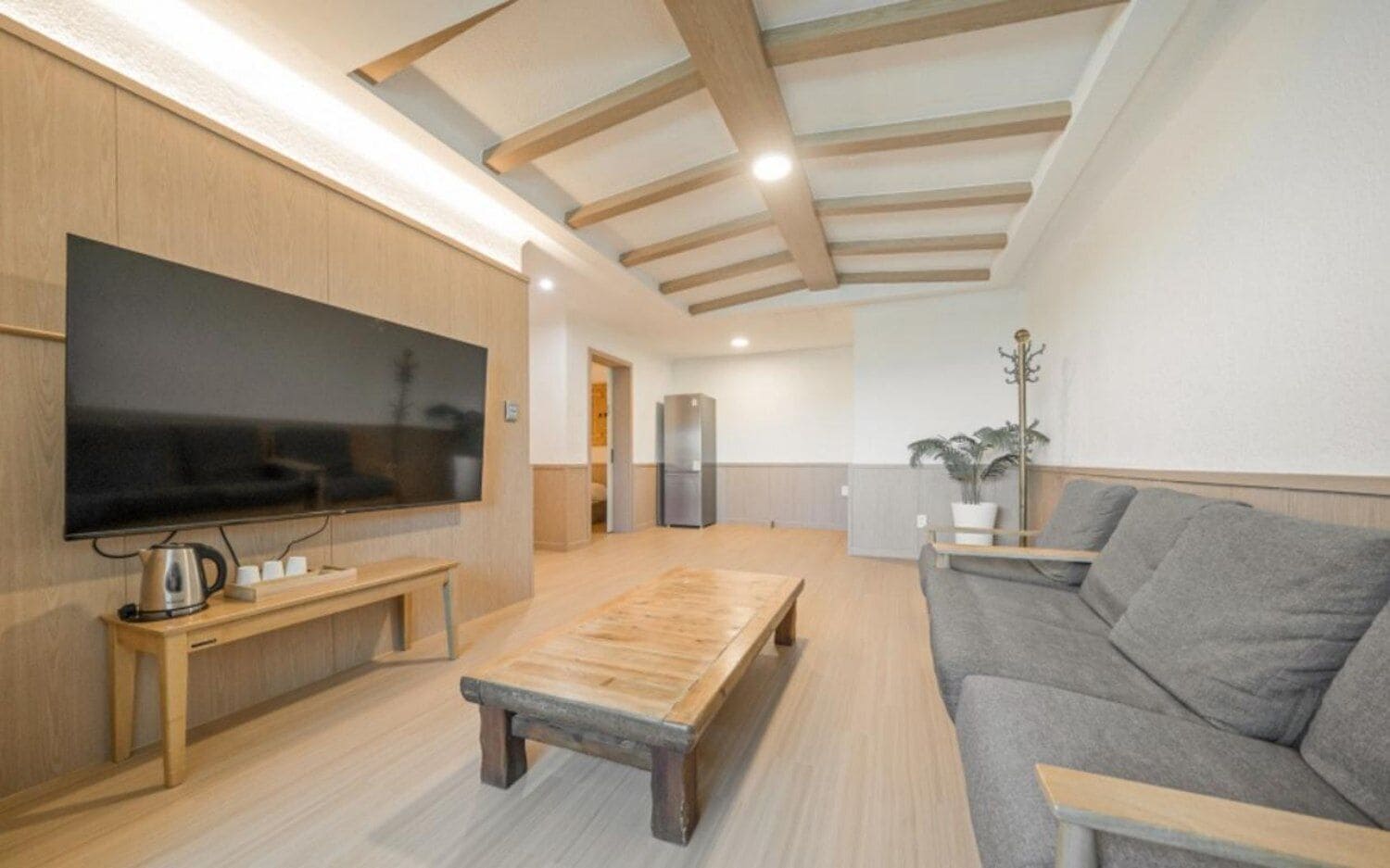 A modern living room with a flat-screen TV, wooden coffee table, grey sofa, and a potted plant.