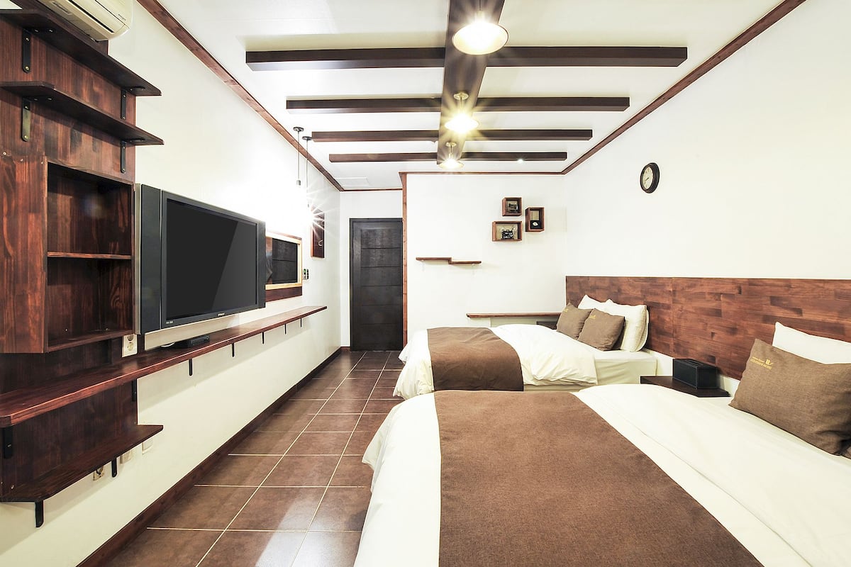 A hotel room with a TV, wooden shelving, a bed with pillows, a bedside table, and a clock on the wall.
