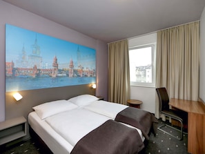 Habitació d'hotel amb un llit gran, una taula de treball, una cadira i una finestra amb cortines.