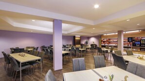 Een ruime eetruimte met meerdere tafels en stoelen, een centrale kolom en een buffet in de achtergrond.