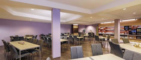 A spacious dining area with multiple tables and chairs, a central column, and a food display area in the background.