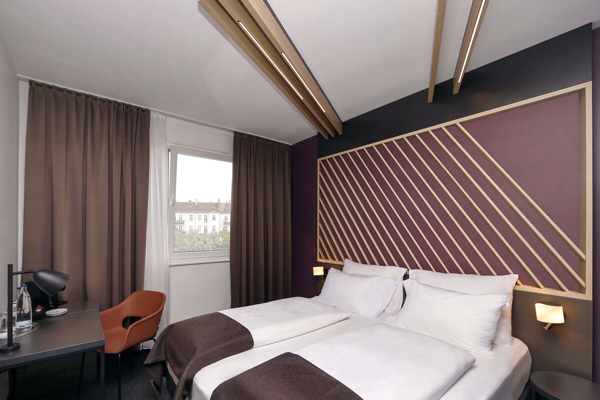 Une chambre d’hôtel avec un lit, un bureau, une chaise et une fenêtre avec des rideaux.