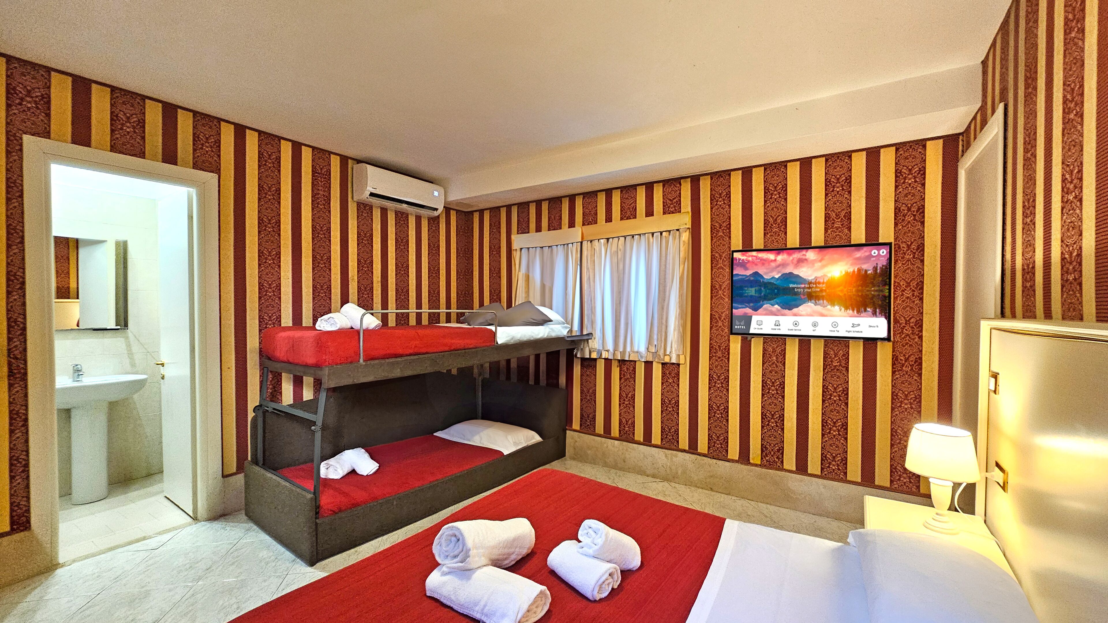 A hotel room with a bunk bed, a TV, a desk with a lamp, and a bathroom with a sink.