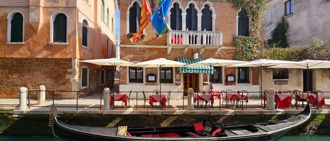 A canal-side restaurant with outdoor seating, a gondola, and historic buildings in the background.