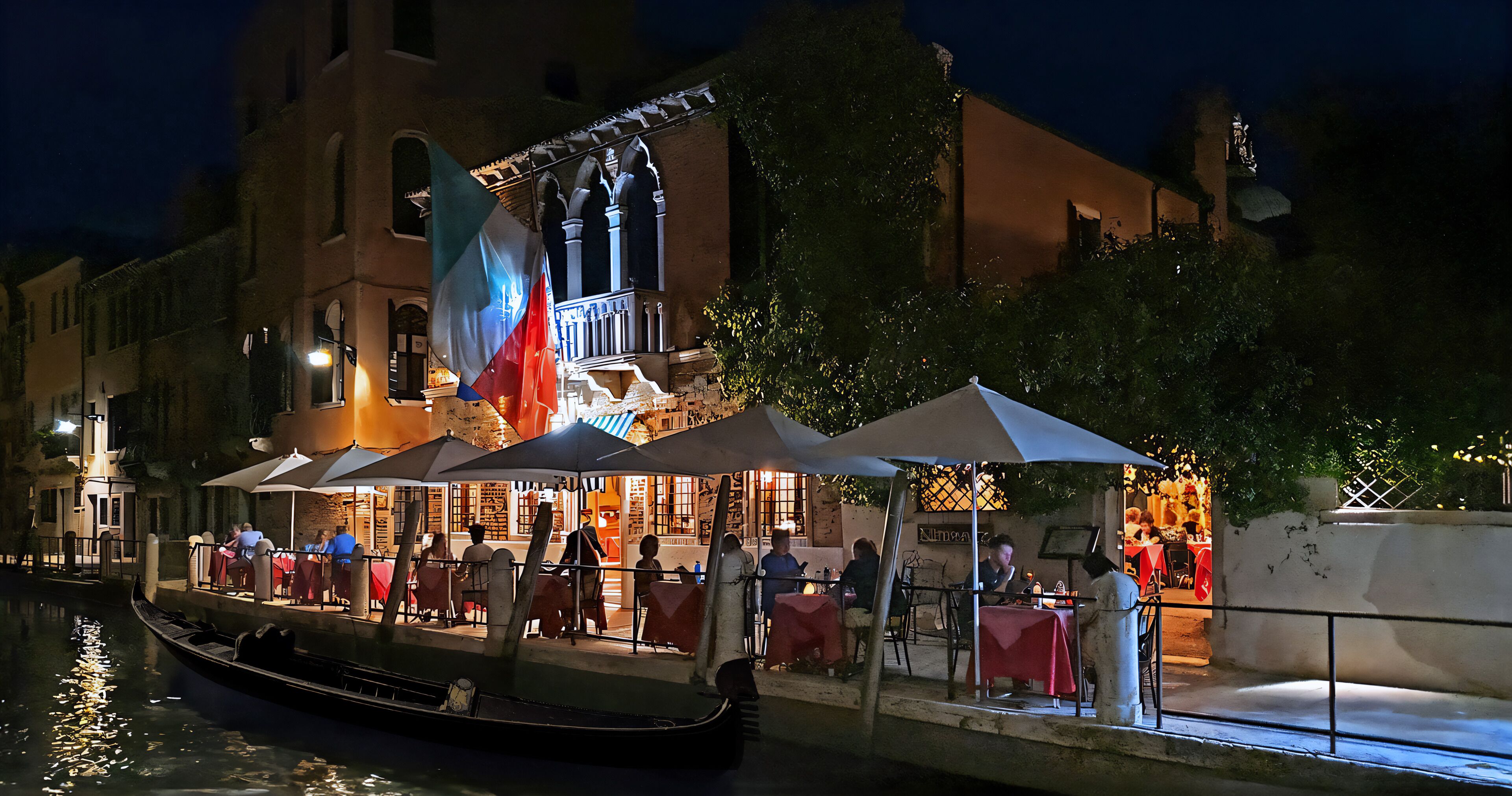 A riverside restaurant with outdoor seating, a gondola, and a flag.