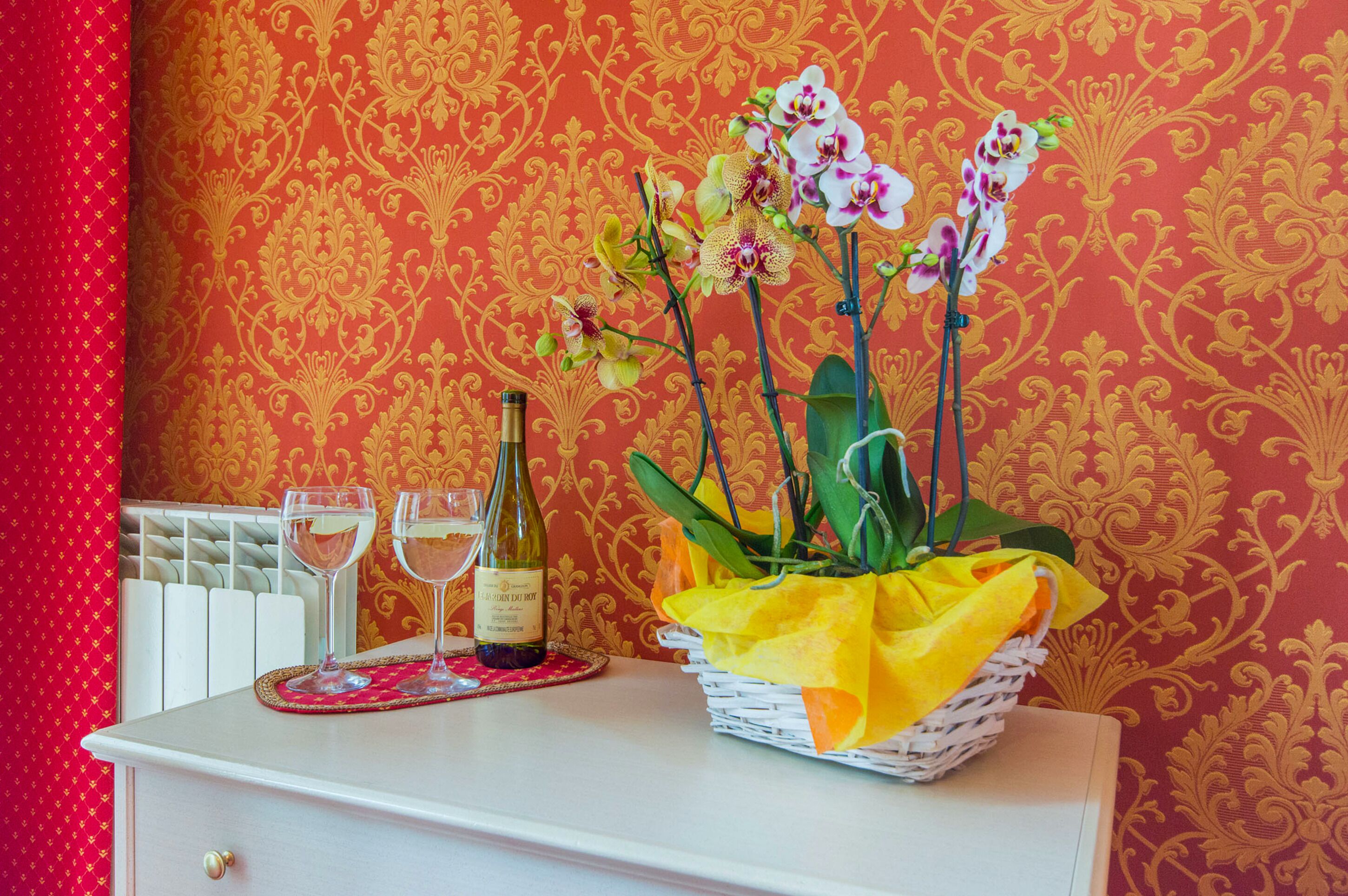 A room with a white dresser, a bottle of wine, two wine glasses, and a basket of orchids.