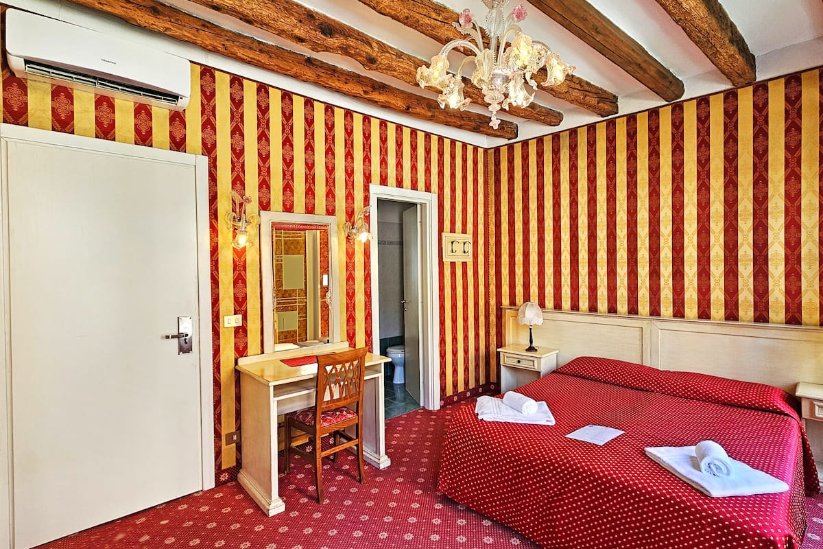 A hotel room with a red and gold striped wallpaper, a wooden bed frame, a white door, a wooden desk with a mirror, a chandelier, and a nightstand with a lamp.