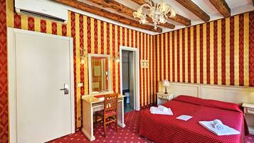 A hotel room with a red and gold striped wallpaper, a wooden bed frame, a white door, a wooden desk with a mirror, a chandelier, and a nightstand with a lamp.