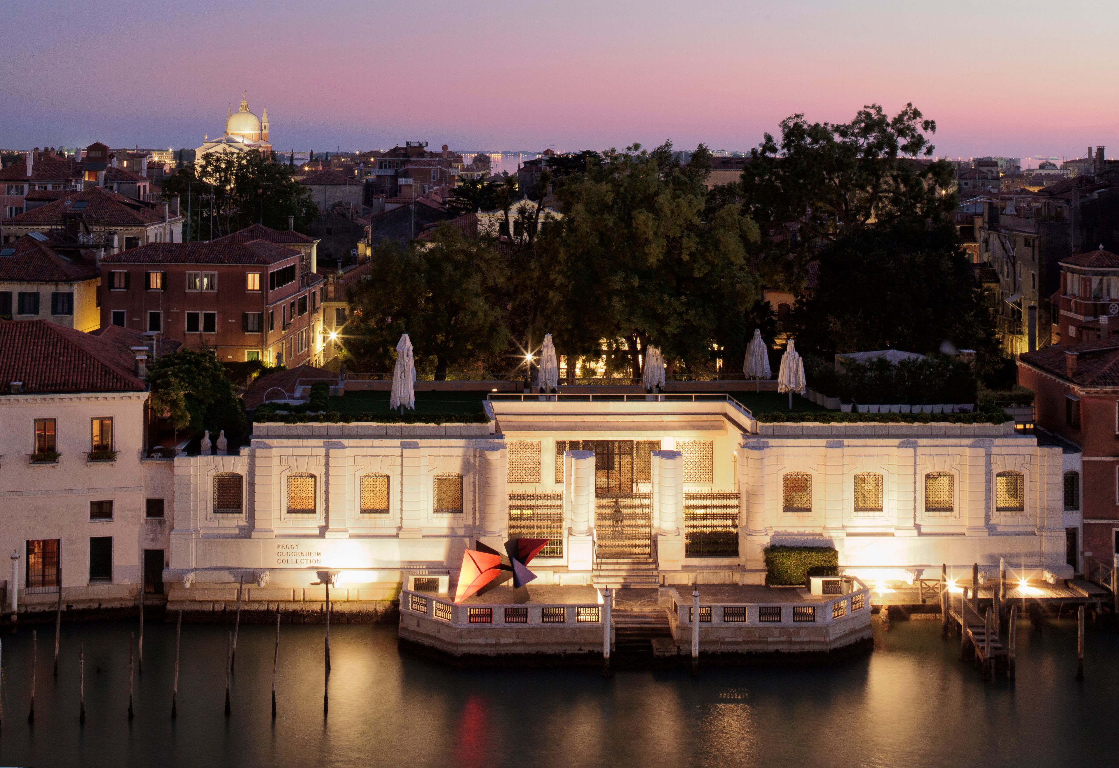 A waterfront building with a central entrance, a domed structure in the background, and a calm river in the foreground.