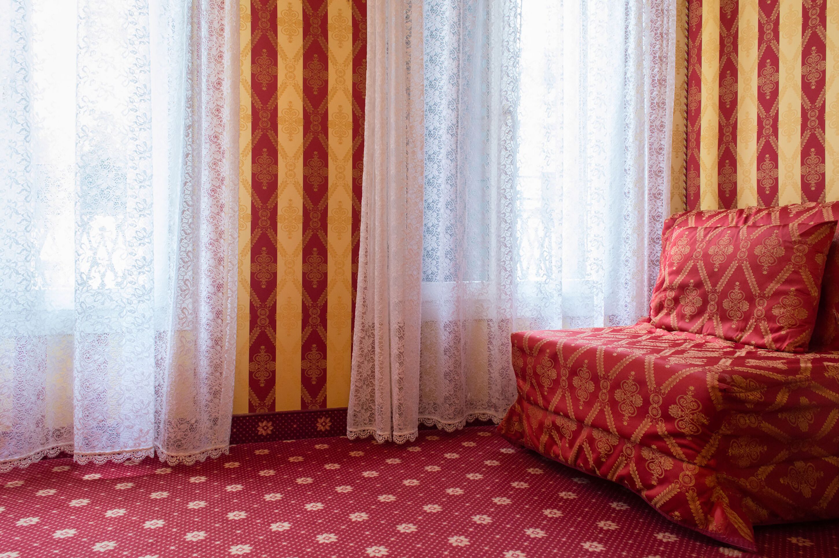 Una habitación con alfombra roja estampada, un sofá rojo y dorado, y una ventana con cortinas a rayas.