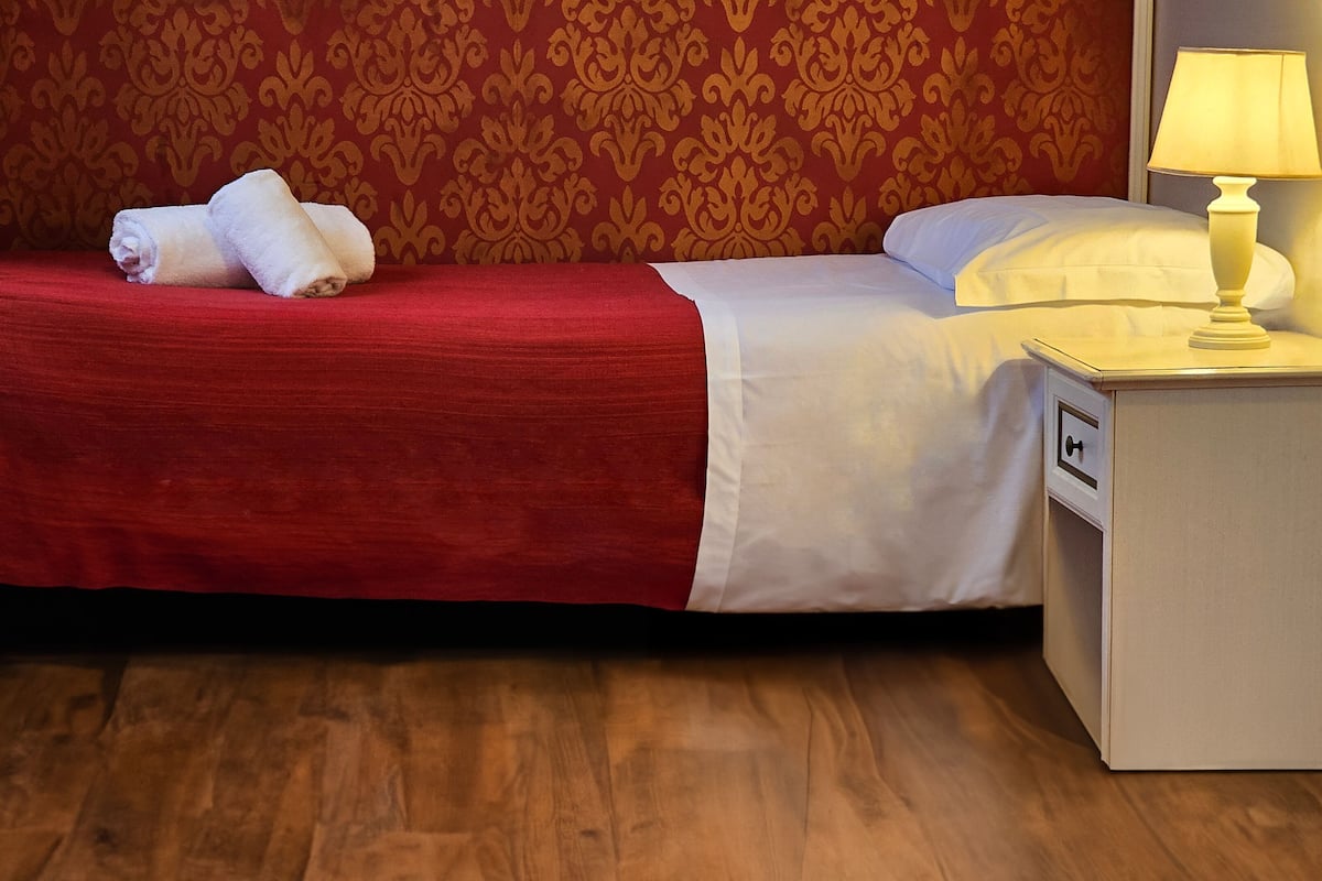 A neatly made bed with a red bedspread and white pillows, a white nightstand with a lamp, and a wooden floor.