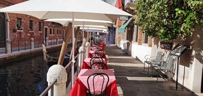 A canal-side restaurant with tables covered by white umbrellas, red tablecloths, and metal chairs.
