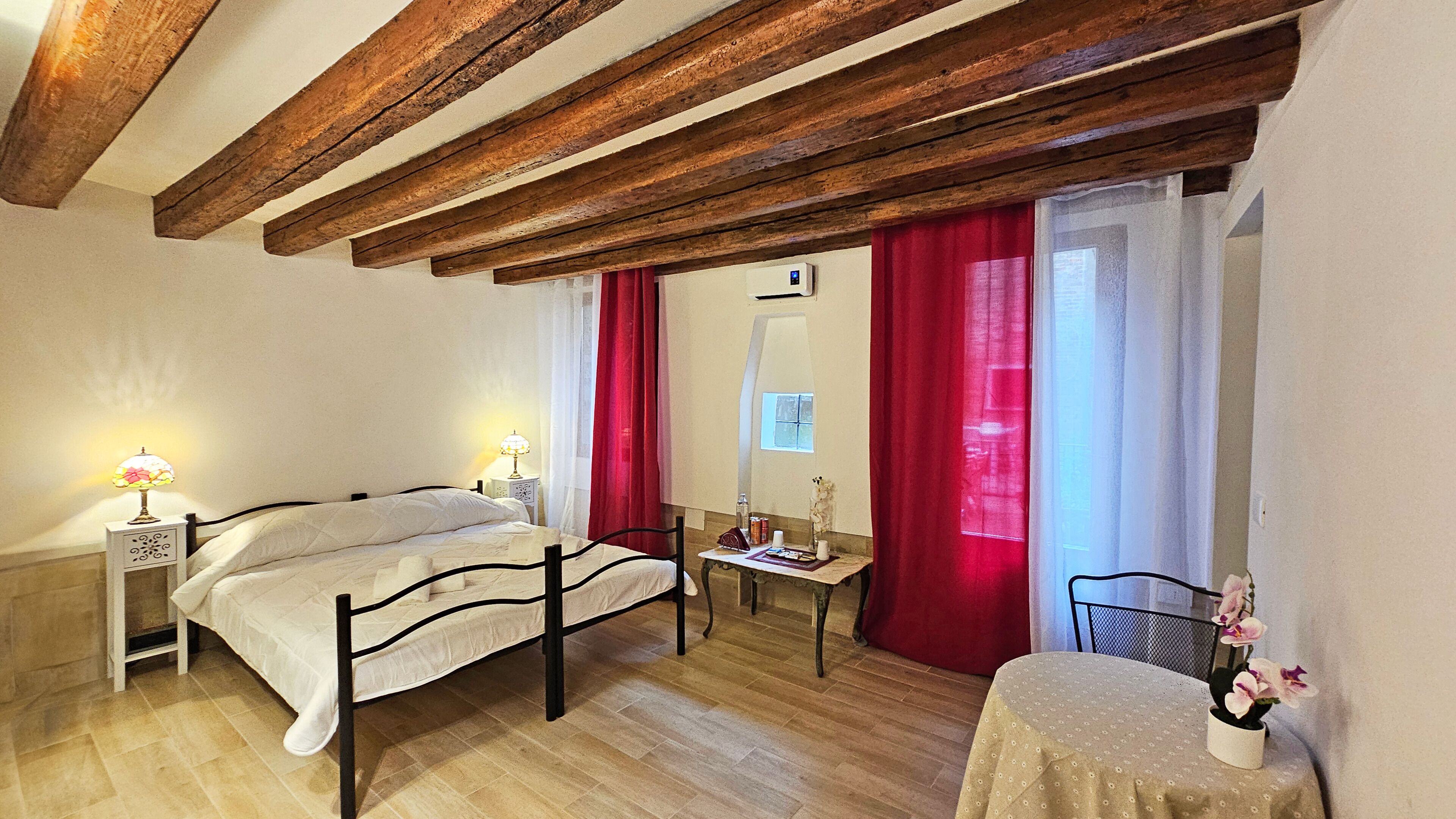 A bedroom with a wooden ceiling, a bed with white bedding, a bedside table with a lamp, a small table with a vase of flowers, and a window with red curtains.
