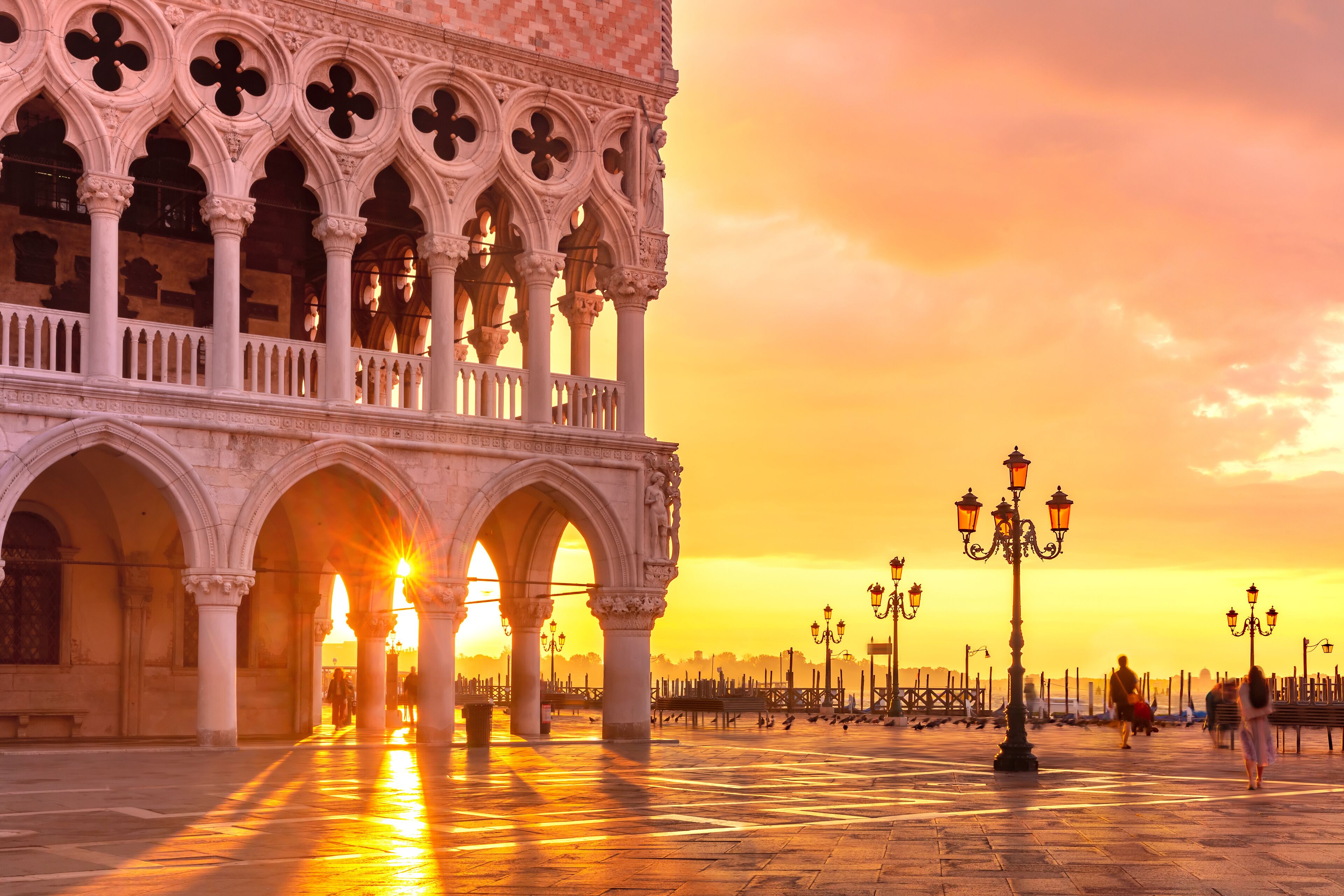 A historic building with ornate arches and columns, a spacious plaza with a fountain, and a sunset in the background.