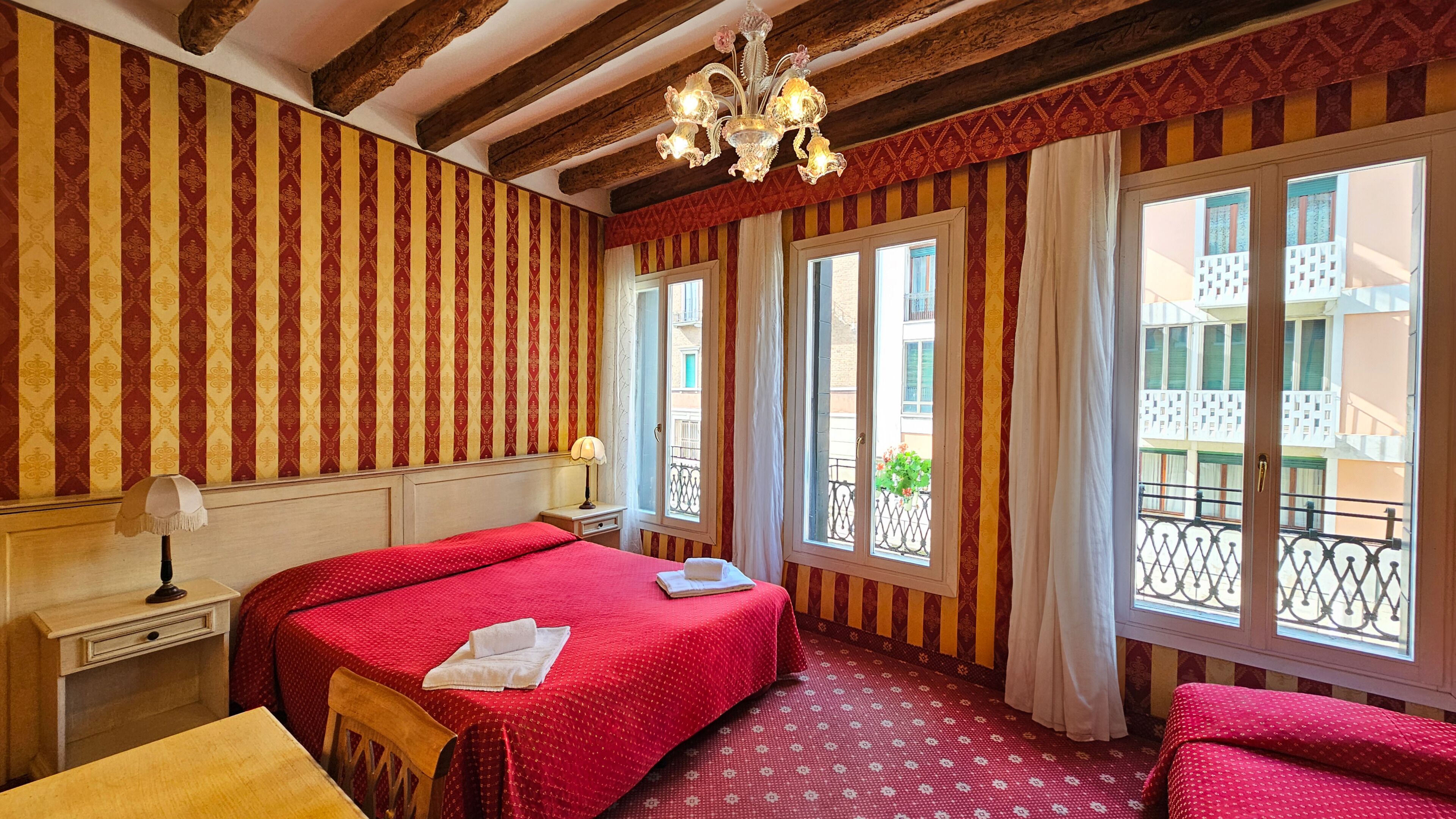 Una camera d'albergo con carta da parati a righe, un lampadario, un letto con biancheria rossa, un comodino e ampie finestre con vista sull'esterno.