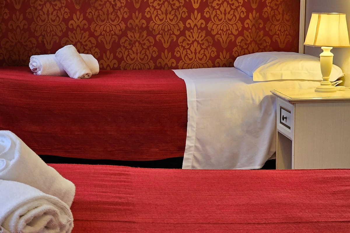 A hotel room with two beds, red and white bedding, a lamp on the nightstand, and a patterned red wall.