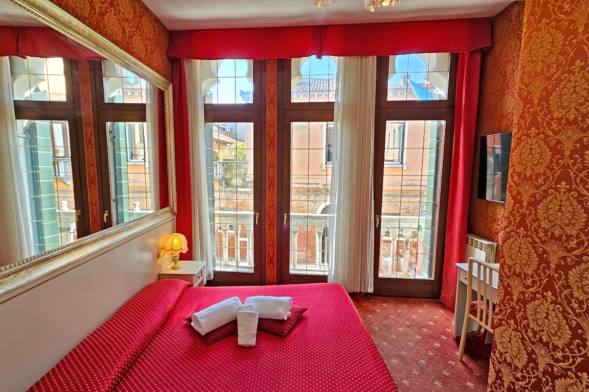 A hotel room with a large bed, a mirror, a TV, and a view of the city through large windows.