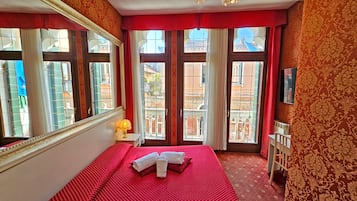 A hotel room with a large bed, a mirror, a TV, and a view of the city through large windows.
