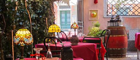 Un espace de restauration en plein air avec des tables recouvertes de nappes rouges, entourées de verdure et illuminé par des lampes décoratives.