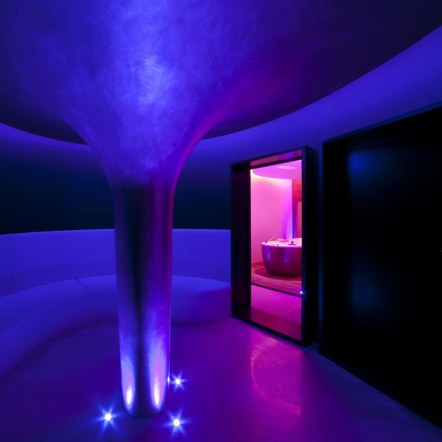 A modern interior with a central pillar, a doorway leading to a room with pink lighting, and a dark wall.