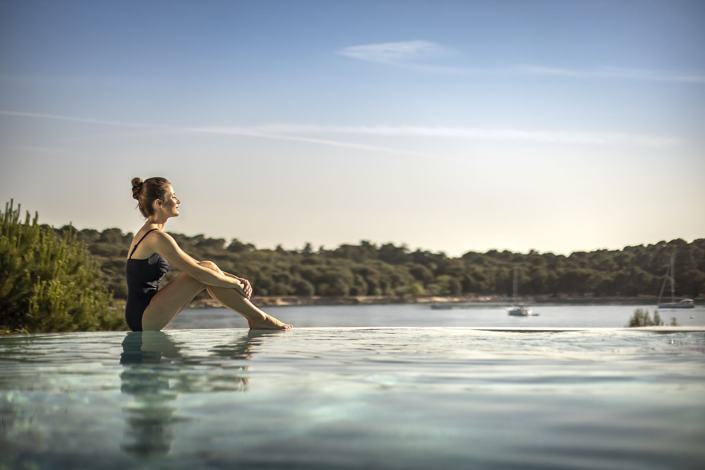 Eine Person sitzt an einem klaren Pool mit Blick auf bewaldetes Gelände und Boote in der Ferne.