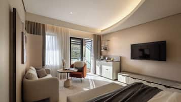 A modern hotel room with a sofa, a small table, a chair, a TV, and a kitchen area.
