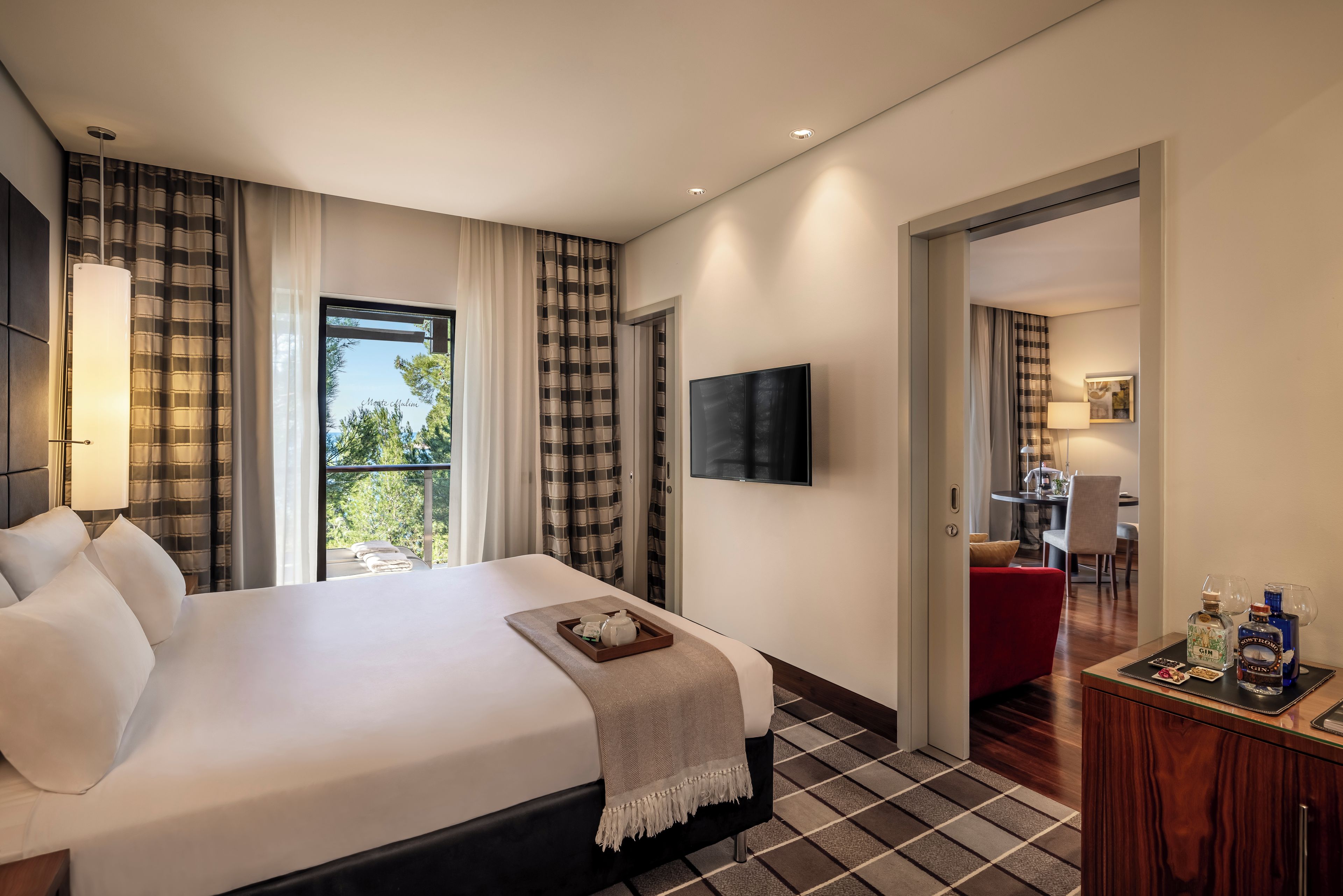 A hotel room with a large bed, a TV, a desk with a chair, and a view of greenery outside.