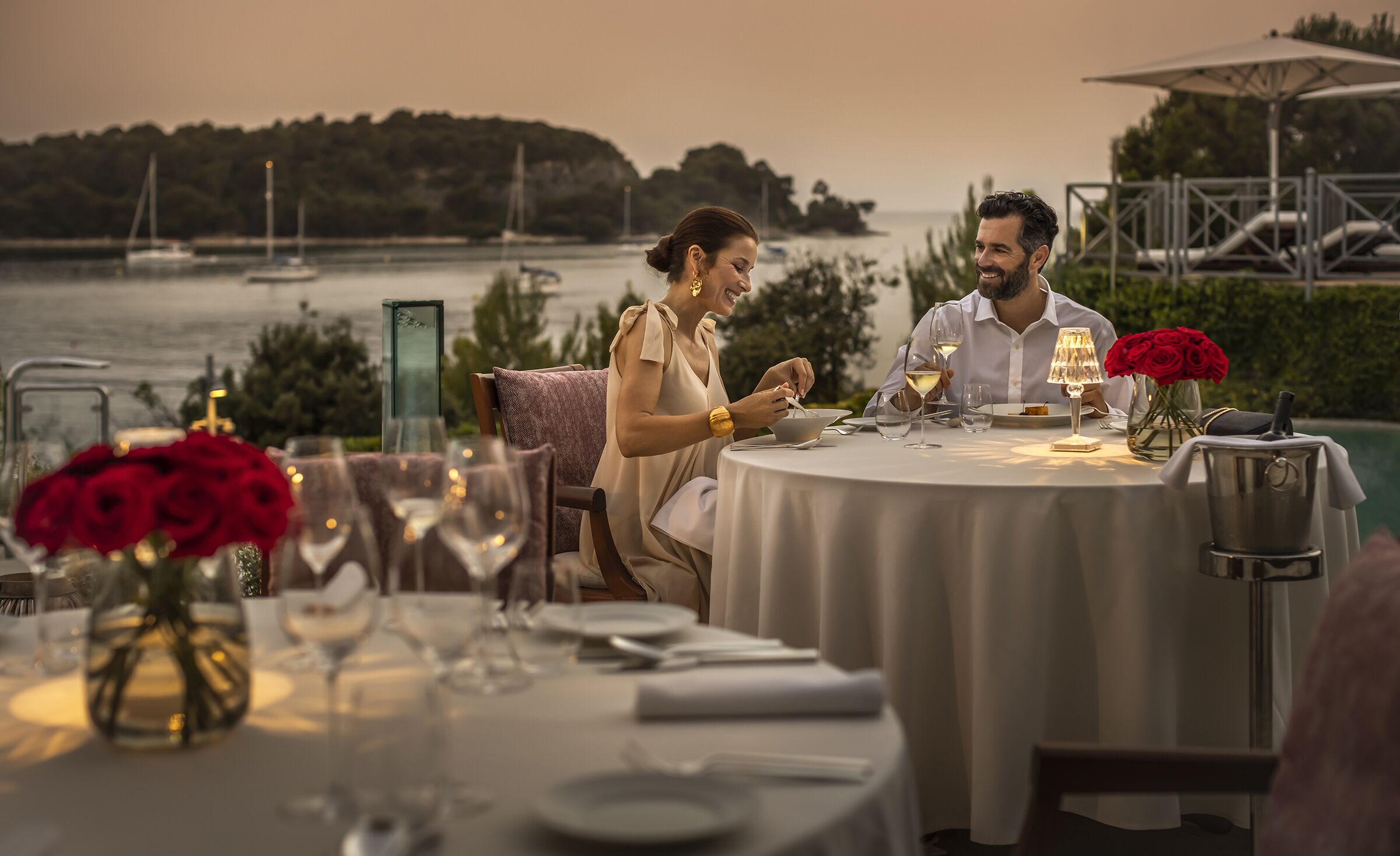 A couple dining at an outdoor table with a view of the sea and boats.