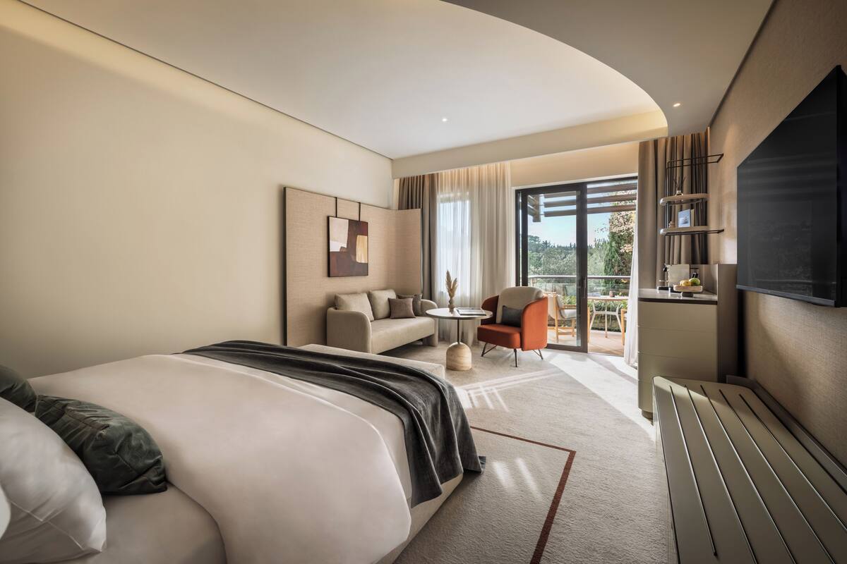 Una habitación de hotel moderna con una cama grande, un sofá, una mesita redonda y un televisor montado en la pared.