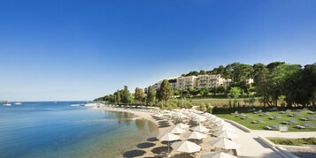 Una località balneare con mare cristallino, spiaggia di sabbia e ombrelloni, e un edificio con numerosi balconi affacciati sul mare.