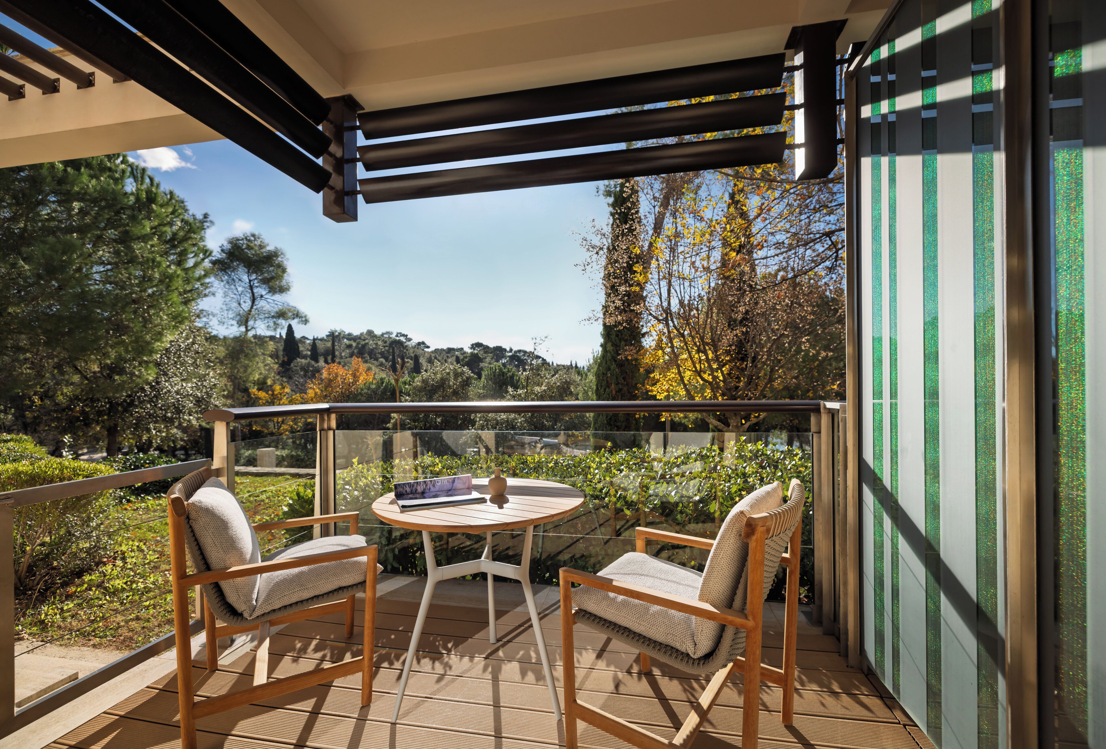 A balcony with two chairs and a table, overlooking a garden with trees and a clear sky.