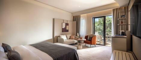 A modern hotel room with a large bed, a seating area with a small table, and a balcony with a view of trees.