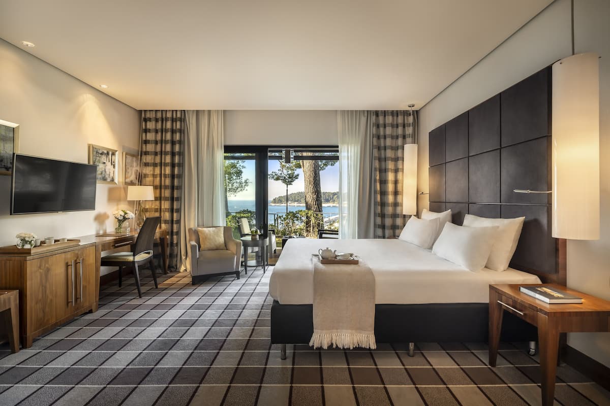 Ein Hotelzimmer mit einem großen Bett, einem Schreibtisch, einem Sessel, einem Fernseher und Meerblick.