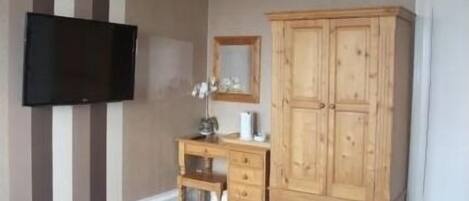 Un dormitorio con cama, armario de madera, escritorio con silla, televisor de pantalla plana en la pared y espejo sobre el escritorio.