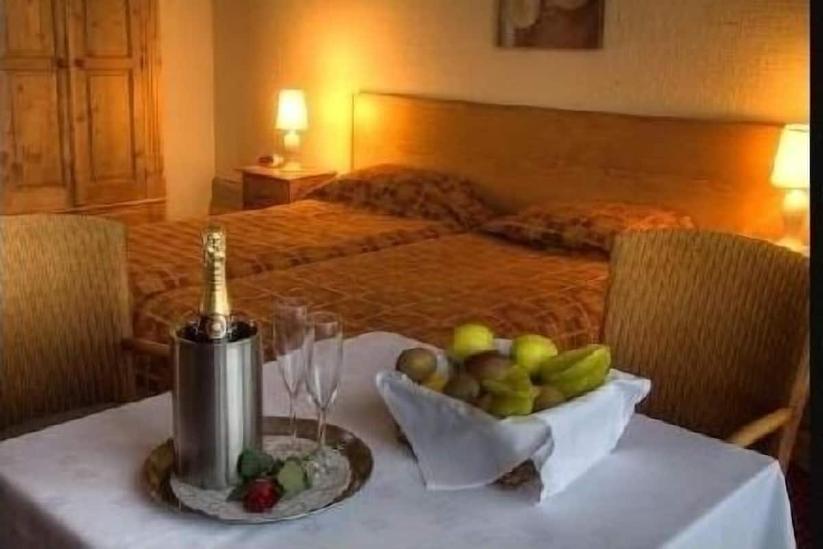 A hotel room with a bed, a bedside table with a lamp, a bottle of champagne, glasses, and a bowl of fruit.