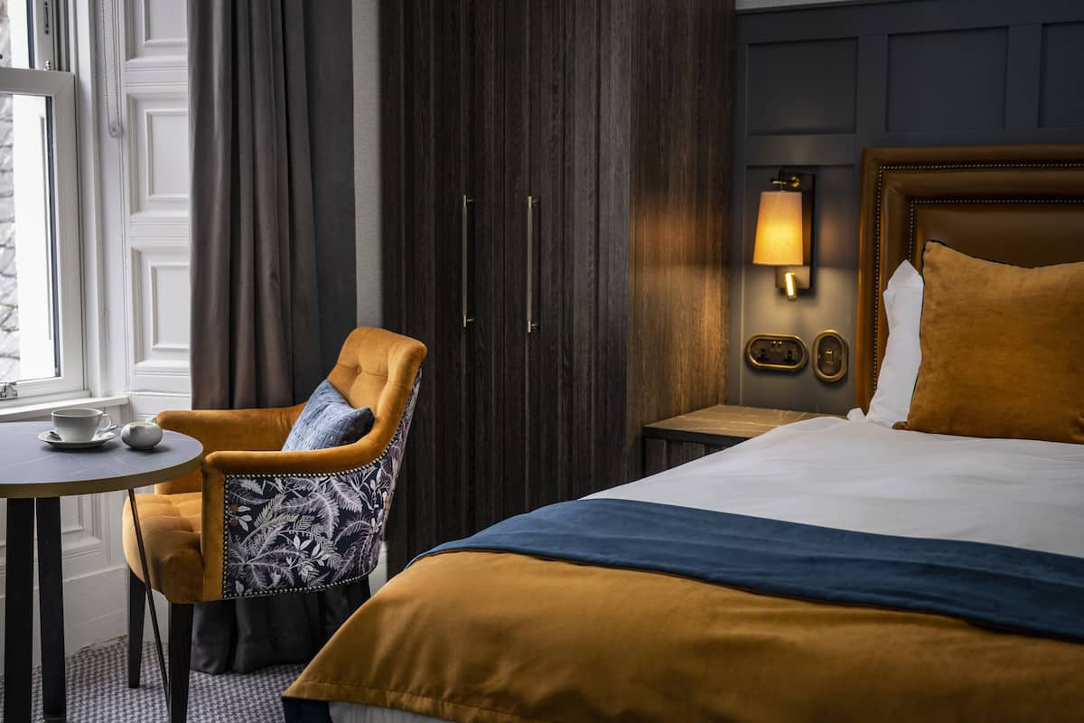 A hotel room with a bed, a chair, a table with a cup and saucer, and a wall-mounted lamp.