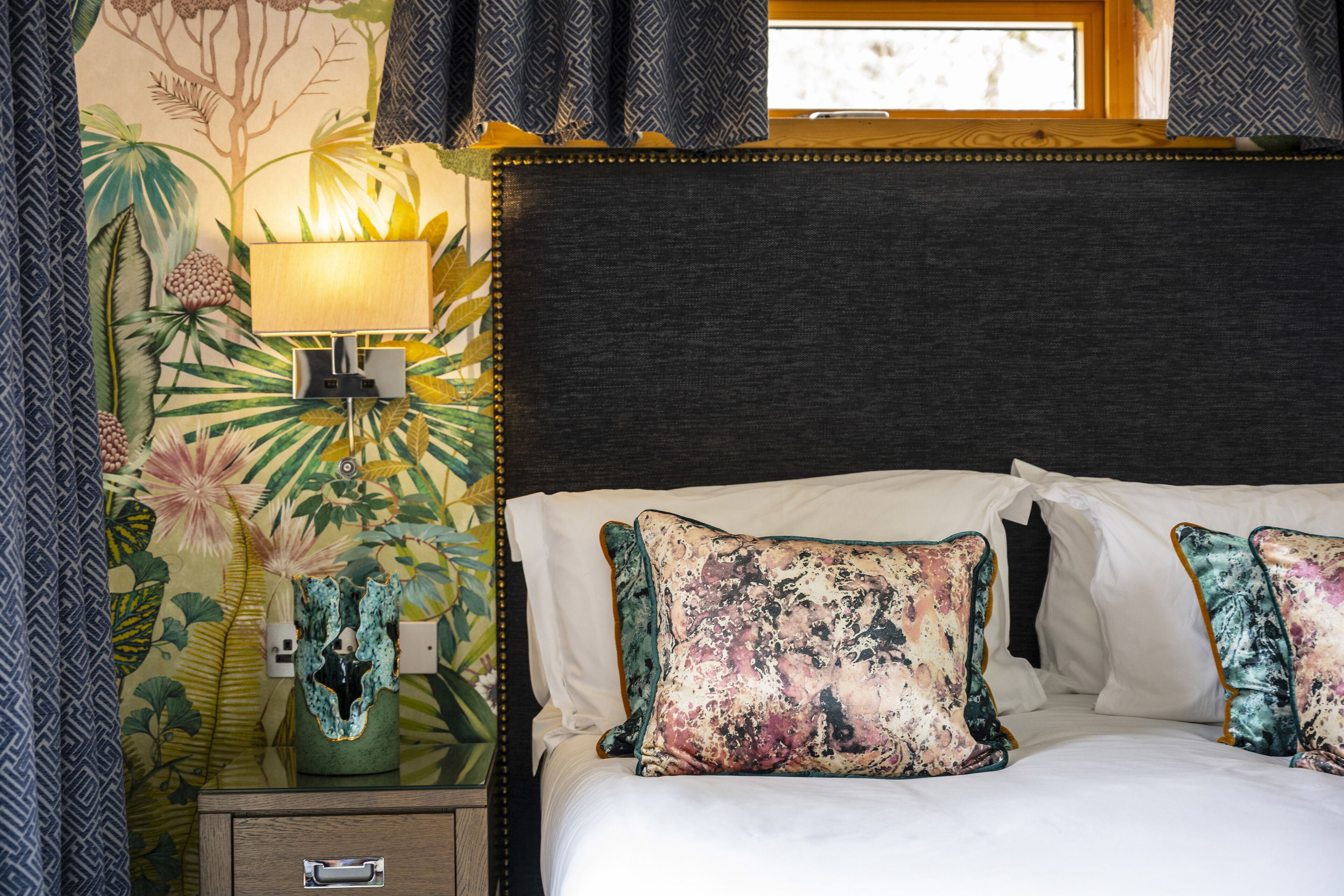 A bedroom with a bed, pillows, a nightstand, a lamp, and a wall with a tropical design.