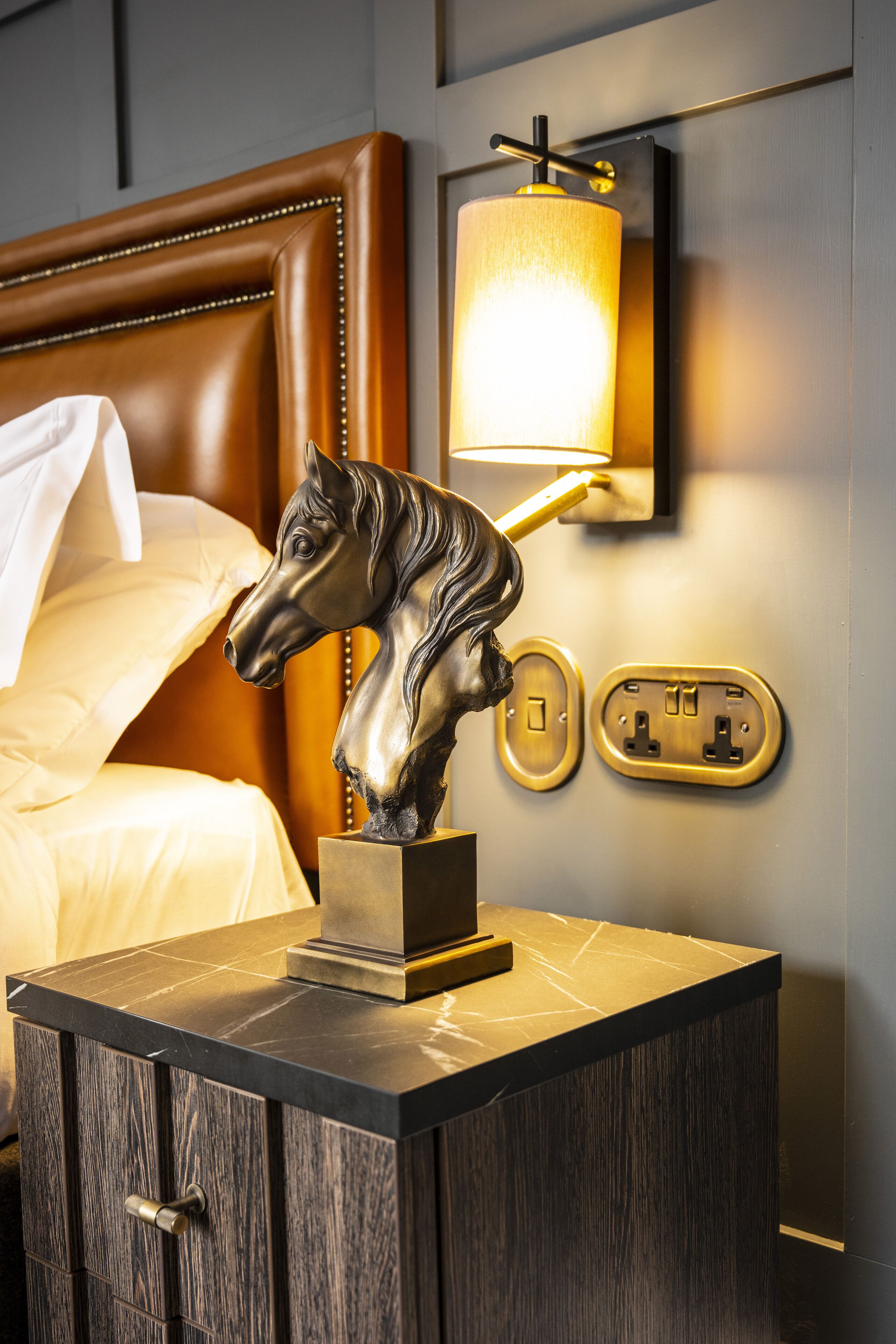 A bed with a headboard, a nightstand with a horse statue, a wall-mounted lamp, and an electrical outlet.