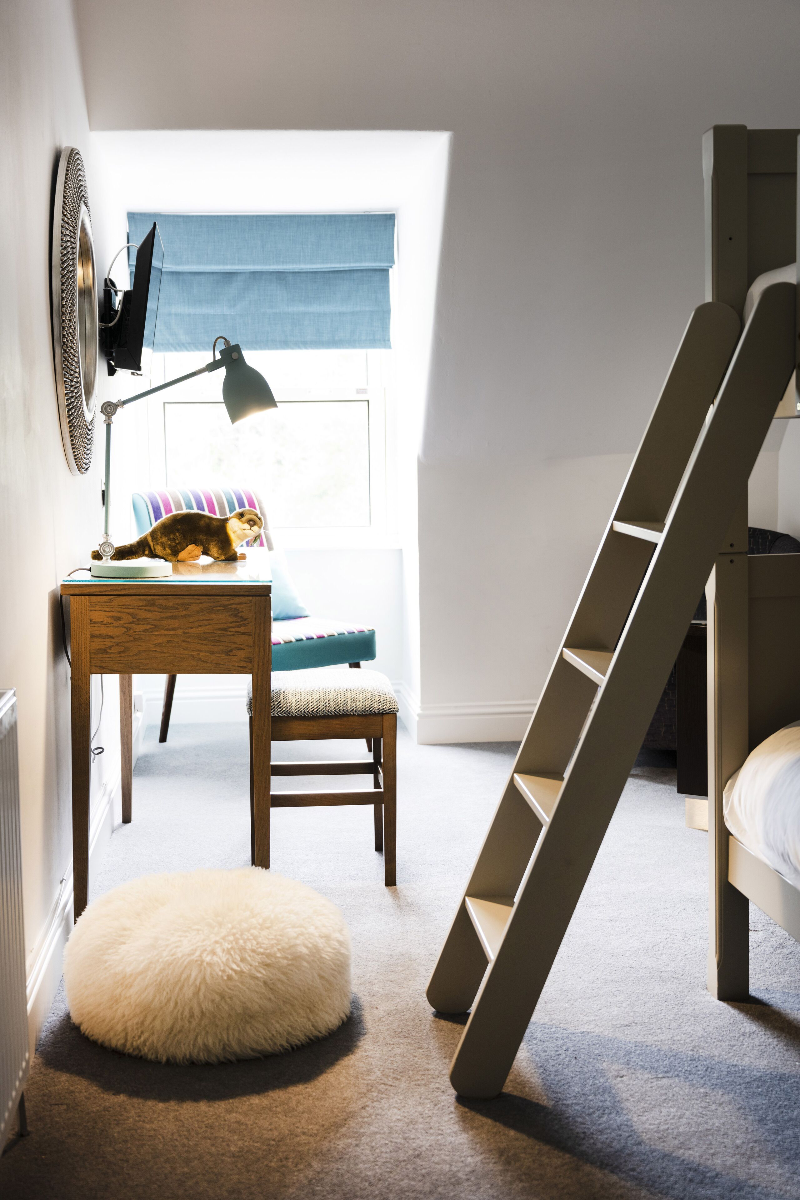 Un dormitorio con una cama nido, un escritorio con lámpara, una silla, un otoman redondo y una ventana con persianas azules.