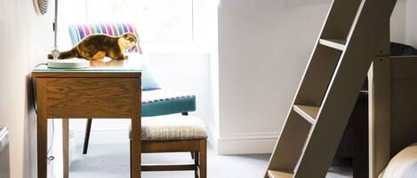 A bedroom with a bunk bed, a desk with a lamp, a chair, a round ottoman, and a window with blue blinds.